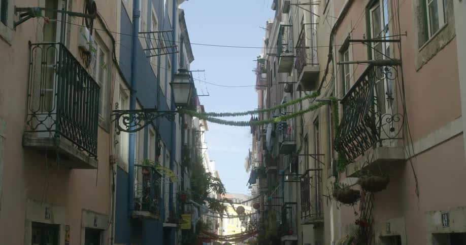 A narrow cobbled street lined with colorful buildings and balconies with the rooftops of a cityscape in the distance