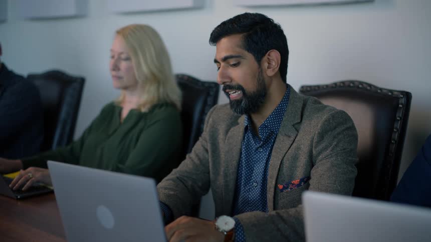 People sitting at a table working on laptop and a bearded man talks