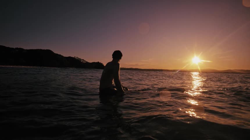 Man sitting on surfboard in the sea with the sun on the horizon