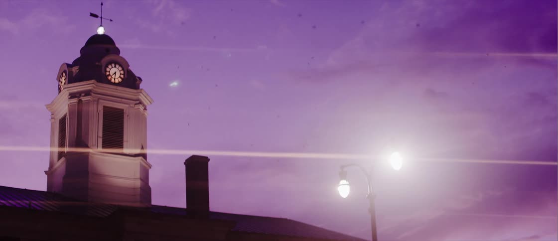 A building with a clock on top and on the right side a street lamp is lit and it is getting dark