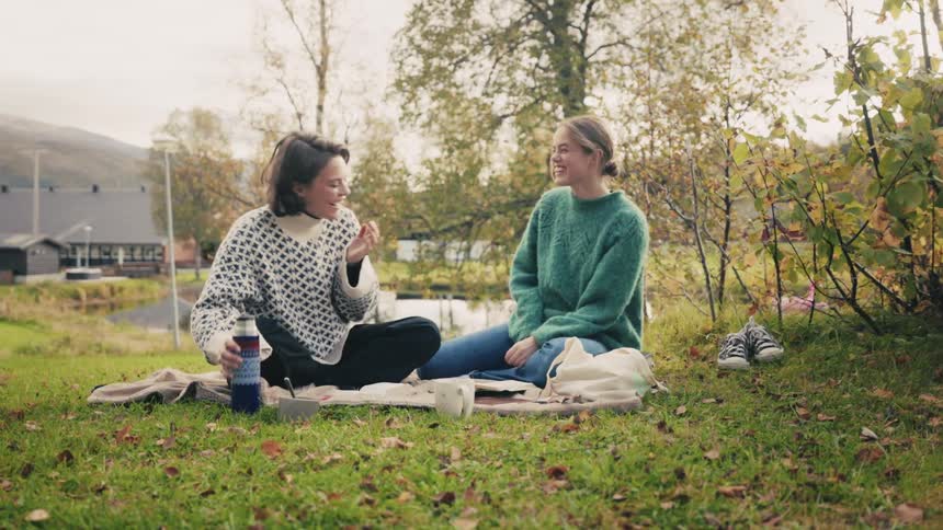 Two women having a picnic on the grass next to a lake and one of them is pouring a liquid into a mug and they laugh