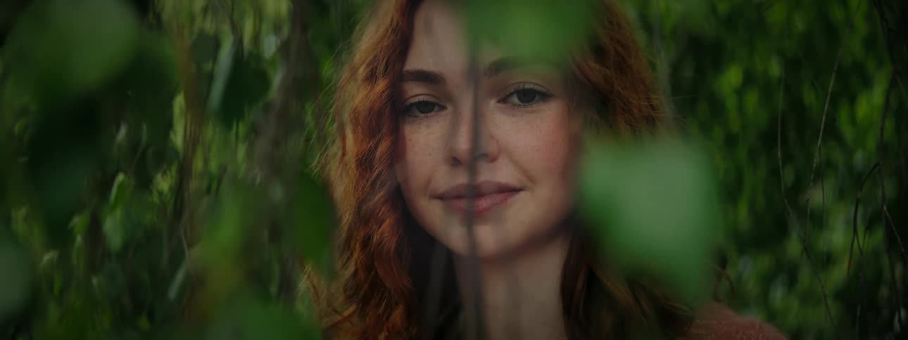 Face of a young redhaired woman hidden among green leaves of a tree