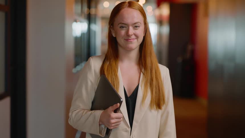 A redhaired woman in a formal outfit holding a laptop in her arm is standing looking forward with a subtle smile