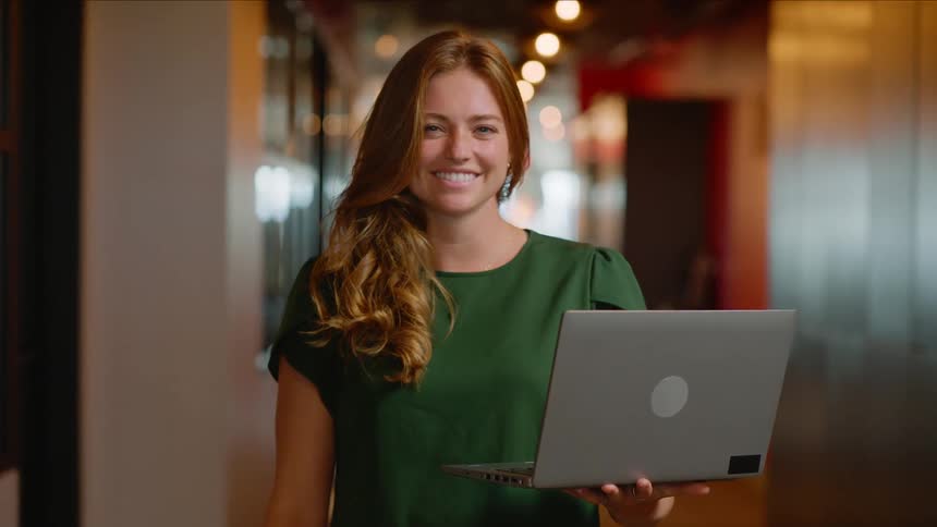 A young woman with long hair smiling at the camera while holding a laptop in a hallway