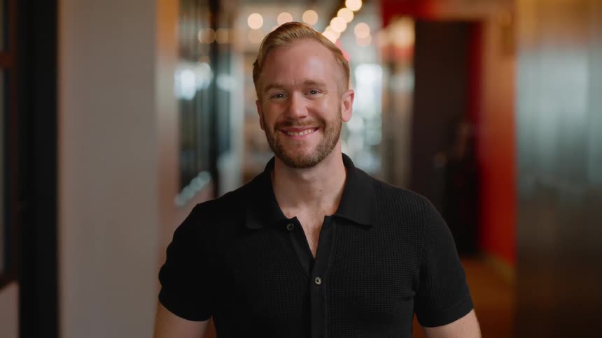 A blonde man in a black shirt is standing looking forward and smiling