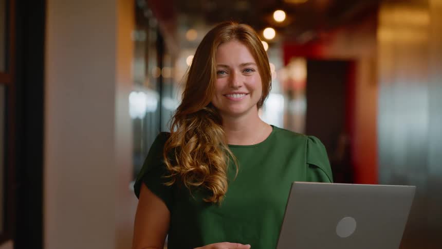 Red haired woman in a hallway giving a smile while holding an open laptop in her hands