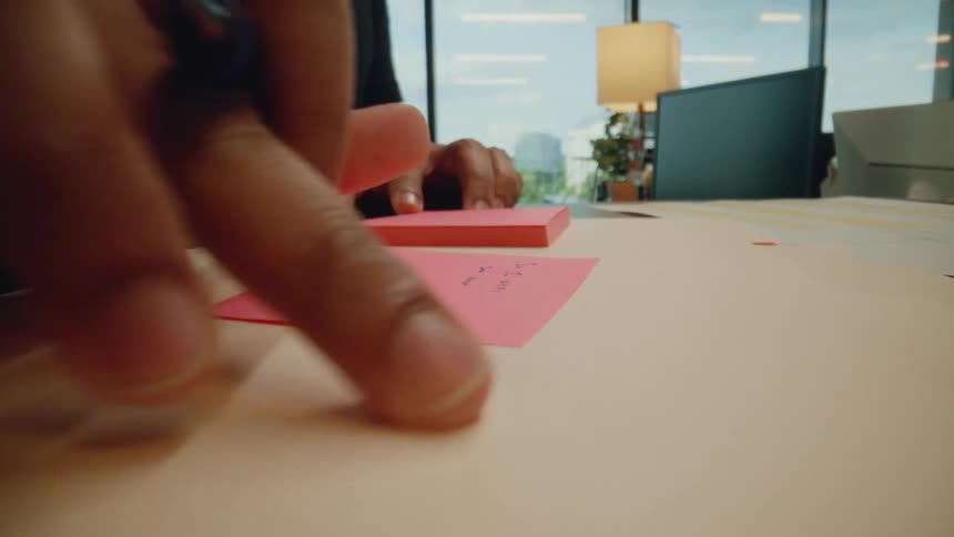 A person writes on a pink sticky note while working at a desk
