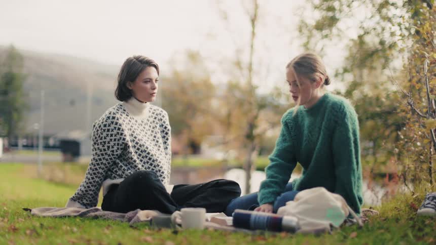 Two young women sitting on a rug in the grass talking