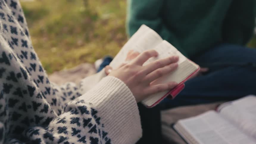 Two people sitting in a lawn reading books one holding up a small one the other reading a large one from the ground