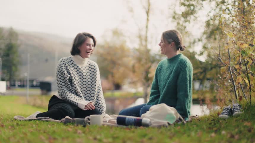 Two women sitting on a blanket on a grass are smiling while talking