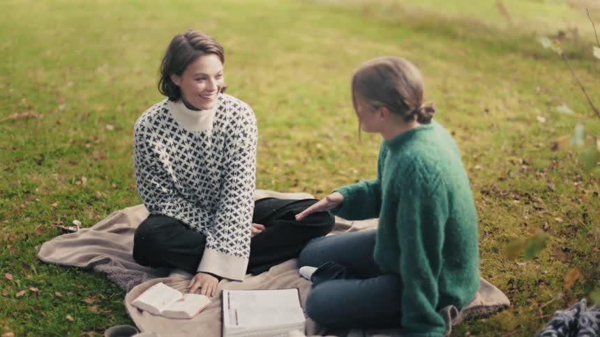 Two women sitting on a blanket on a grass and they are holding open books and smiling at each other
