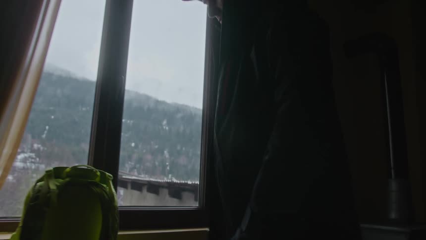 A man by a window with a mountainous landscape view outside putting on a jacket