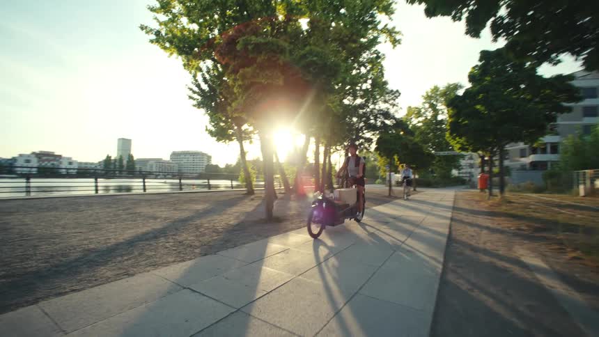 A woman rides a long bicycle along a path next to a river in a city