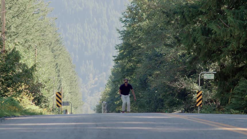 A man is skateboarding on the road with his dog