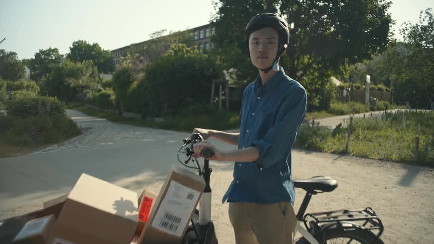 A delivery boy wearing a helmet a blue shirt and brown pants poses with his cargo bike loaded with boxes