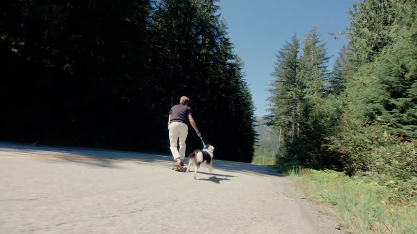 Man wearing sunglasses a blue shirt and white pants rides a skateboard down the road with his dog in the forested area