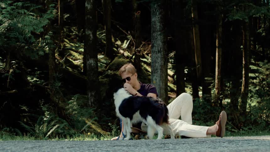 A man wearing sunglasses sits outdoors next to a path while petting a dog