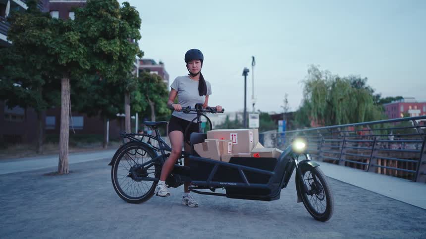 A woman wearing a helmet standing next to a cargo bike loaded with boxes on a paved path surrounded by trees and urban structures in what appears to be the evening