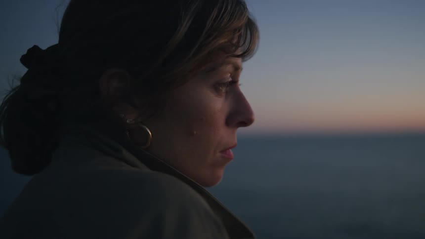 A woman wearing a coat stands outside as the sun is low in the sky