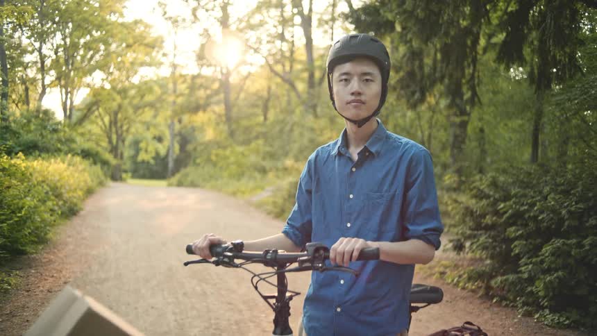 A delivery boy wearing a helmet a blue shirt and brown pants poses with his cargo bike loaded with boxes on a dirt road in the forest