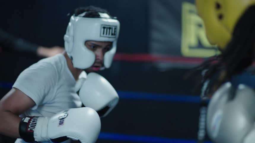 A young boxer wearing white headgear and gloves facing an opponent in a training session with a focused and determined expression