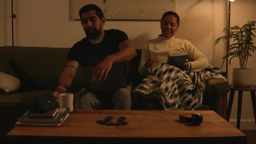 Woman hands a man using a computer a mug then sits down next to him to read a book