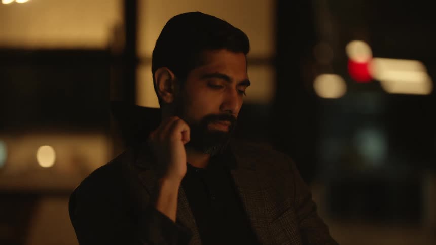 A young bearded man sits waiting with blurred city lights illuminating the night background