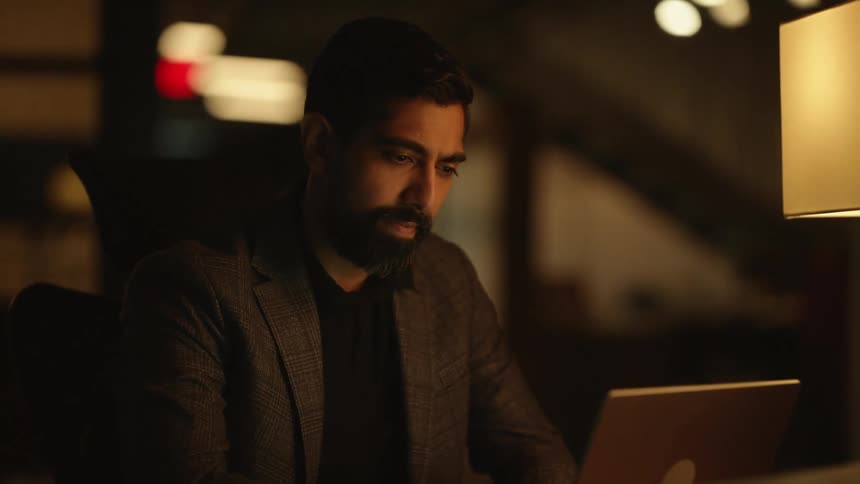 A man with a beard works on his laptop in a dimly lit room
