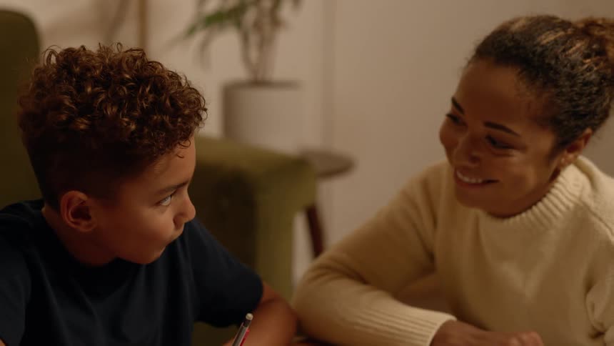 Cheerful mother sitting next to her son who is studying