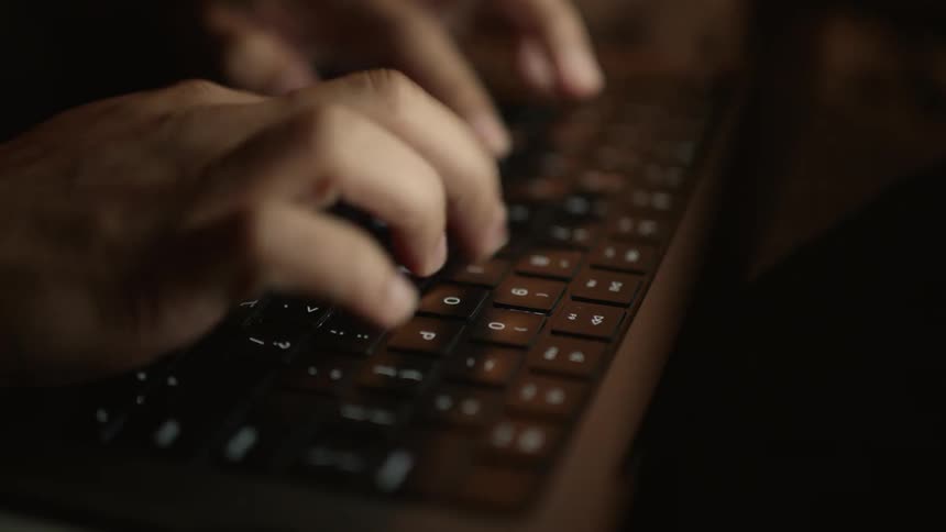 A pair of hands is diligently typing on a computer keyboard in a dimly lit room