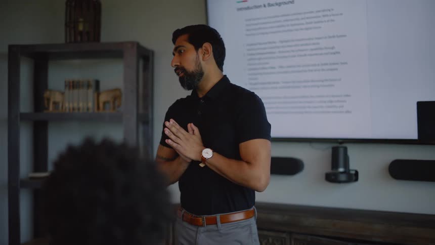 A bearded man in a black shirt presenting to a small group in a meeting room with a large screen displaying a document behind him