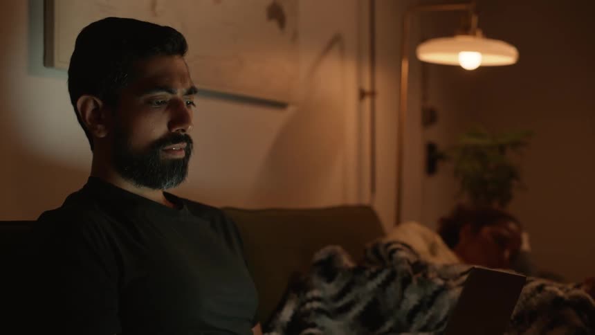 Man sitting interacting with a laptop and a woman lying next to him sleeping on a couch