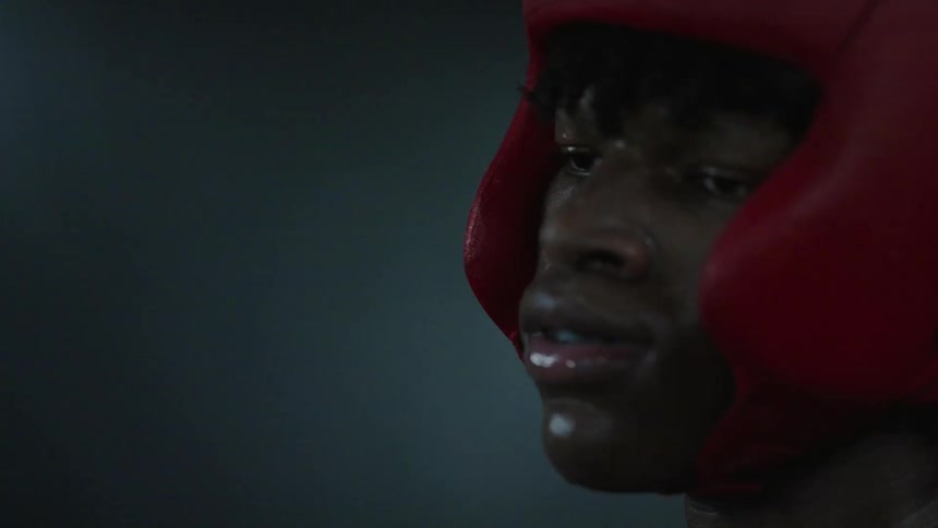 A boxer in red headgear being given water from a plastic bottlebetween rounds with a coach assisting