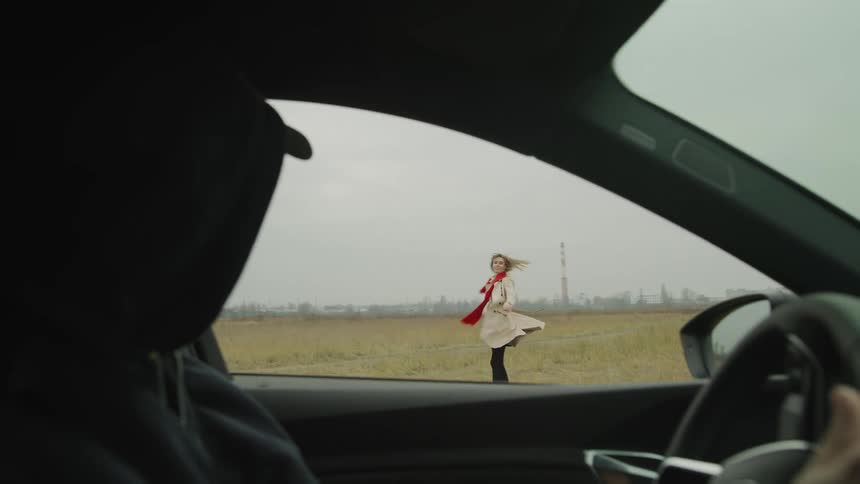 Hooded person inside a car driving and looking at a woman in a field