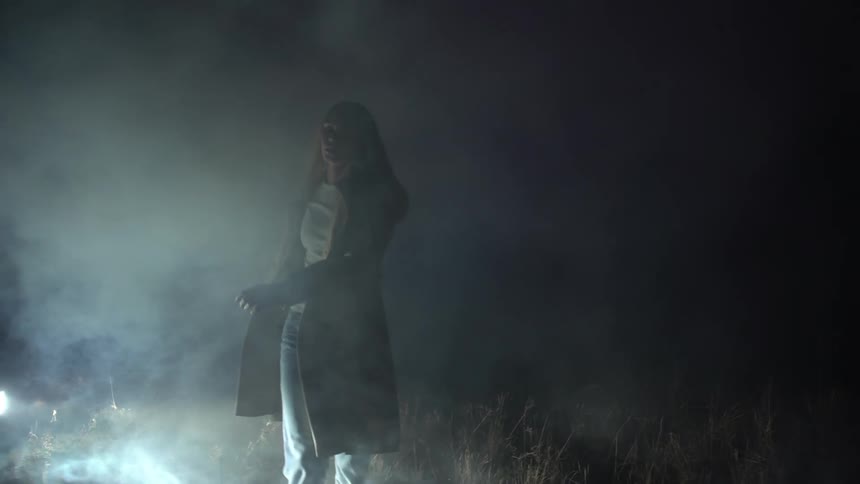Woman walking in a smoky field at night next to a car with its headlights on