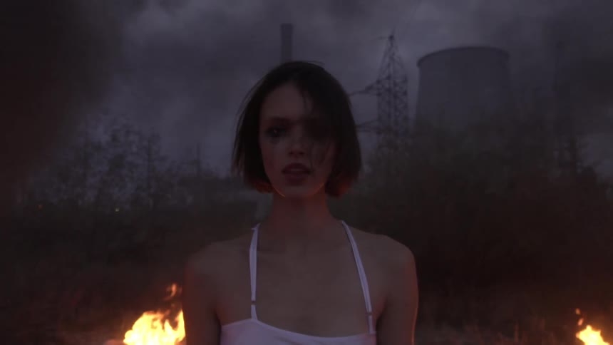 Small fires burn behind a disheveled beautiful young woman surrounded by smoke in an industrial landscape