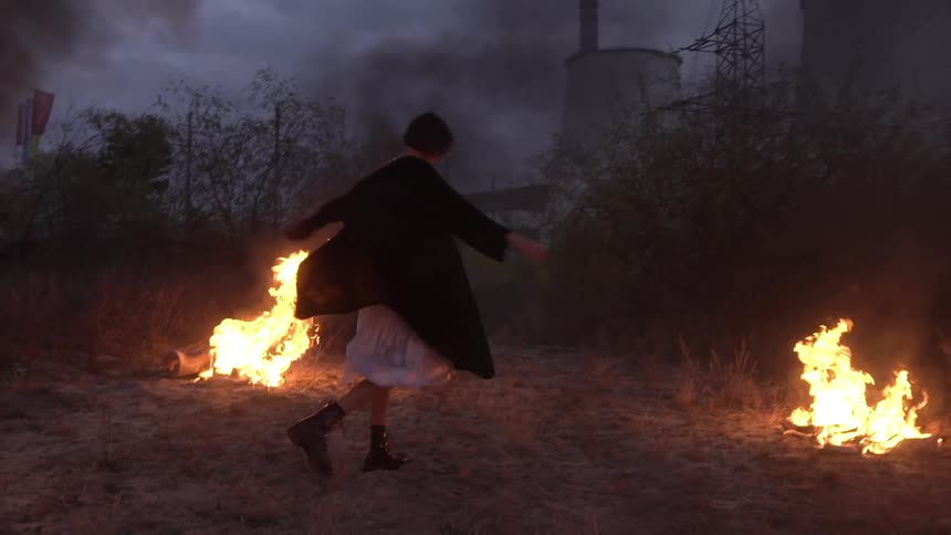 Woman dances in a smokey area among burning fires near a power plant after dusk