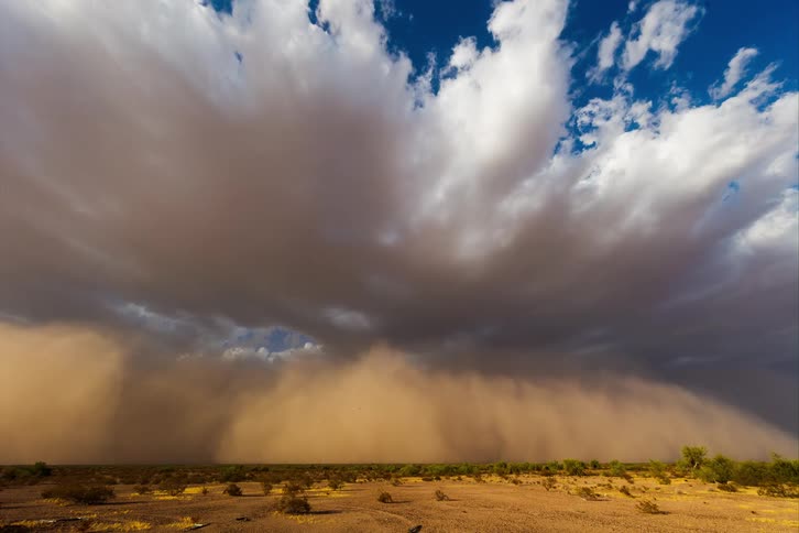A big sandstorm coming quickly in a desert with several white clouds in the sky