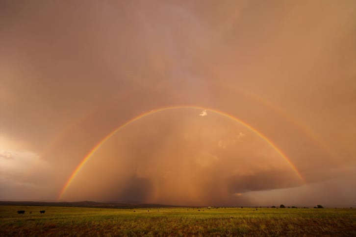 Landscape with a vibrant rainbow arching over a vast field and dark storm clouds in the distance