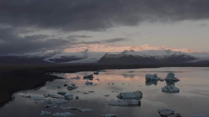 Natural landscape of a lake reflecting the cloudy sky with red hues and pieces of ice with snowy mountains in the background