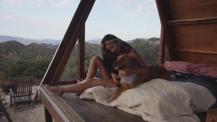 A woman sits on a bed in a small wooden structure in the desert caressing her dog