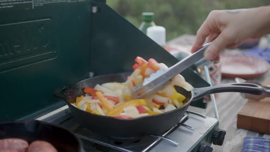 A hand with a tattoo holding tongs turning vegetables on a cast iron skillet on a portable stove