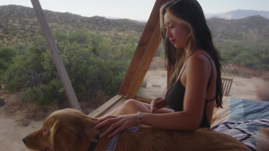 A woman sits on a bed in a small wooden structure in the desert reading a book while her dog lies beside her