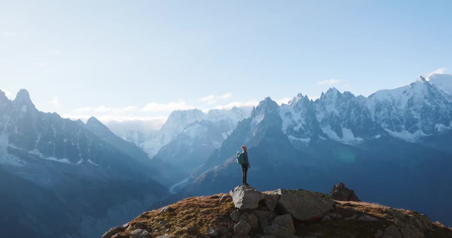 A hiker stands atop a rocky mountainside overlooking a majestic range of snowcapped peaks