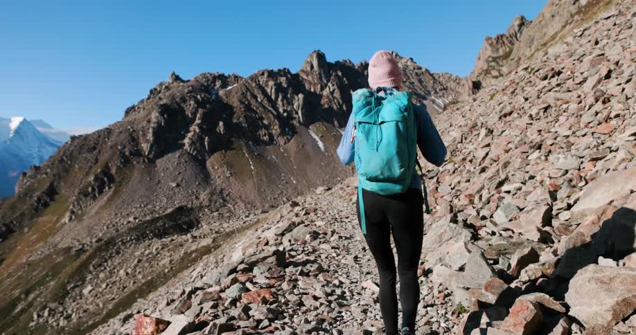 A woman wearing a blue backpack is facing away and walking on a rocky mountain