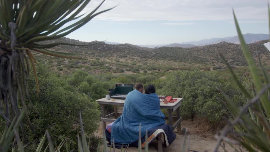 A couple is hugging each other as they sit in front of a wooden table outdoors in a lowlying area and they are wearing a blue blanket around them