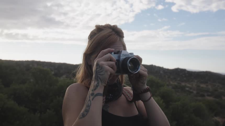 Cheerful woman taking pictures with a vintage camera while standing in an environment surrounded by greenery and hills