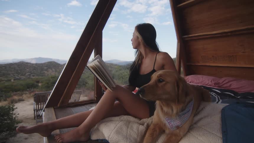 Woman sitting in a camping cabin holding a book with a labrador retriever by her side
