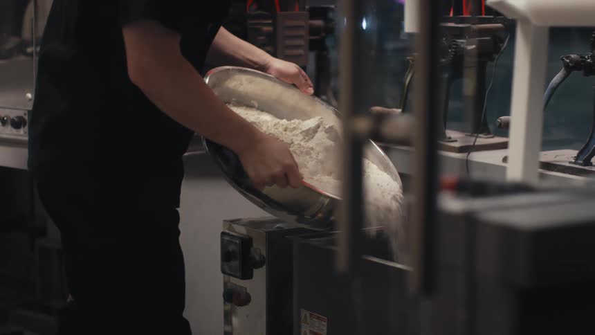A restaurant worker pours a large bowl of flour into a metal basin