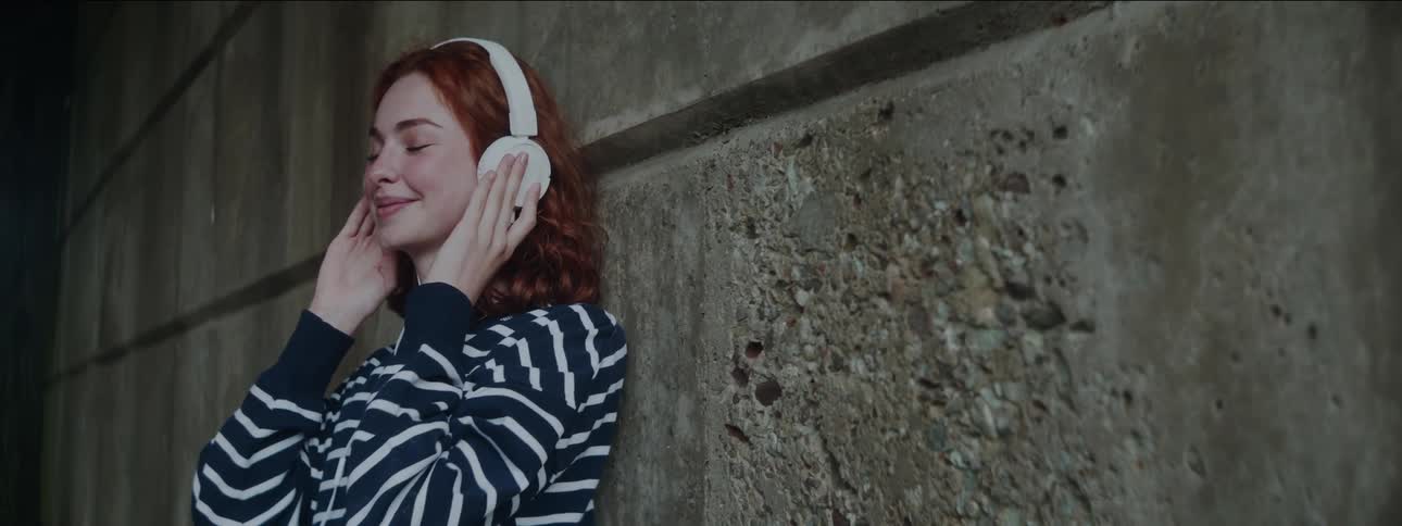 Woman leaning against an exterior building wall listens to music on headphones